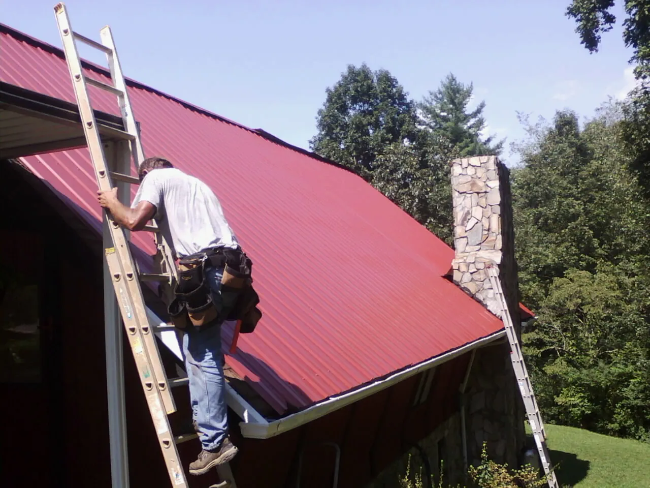Roofer on a ladder beside a red metal roof and stone chimney — Soffit & Fascia Repair by Roofing Brothers in Offutt Afb